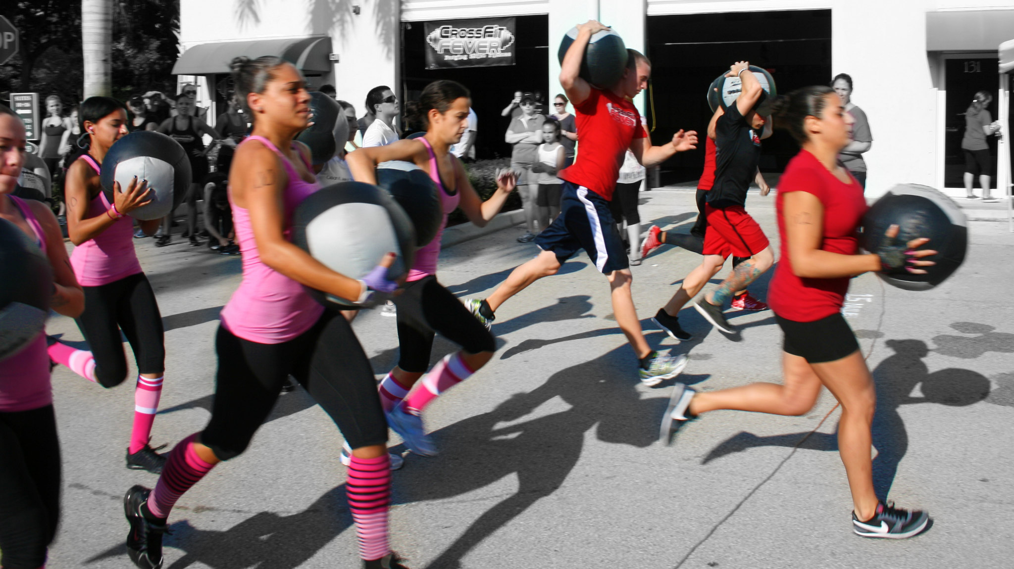 In the foreground, a woman with a pink shirt runs while carrying a medicine ball. In the background, other women and men also run with medicine balls.