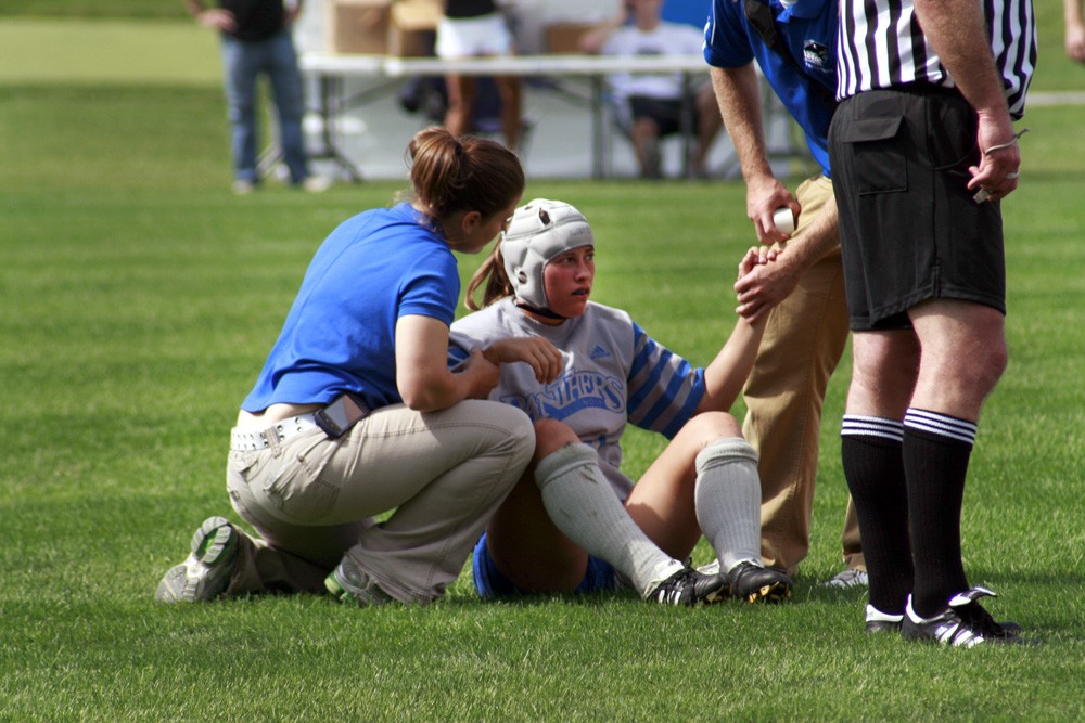 A rugby player sits on the grass, while two athletic trainers attend to her. The trainers hold her hands and appear to be ready to help her to her feet.