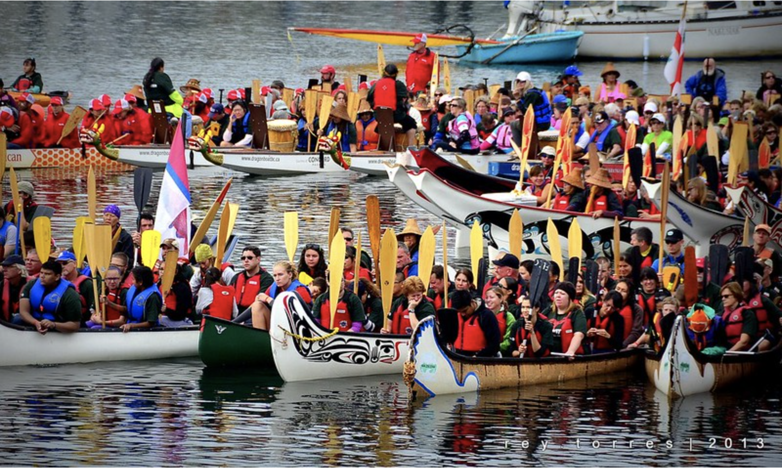 Approximately a dozen colorful dragon boats and traditional Indigenous canoes float on the wather. Each boat is filled with people holding oars.